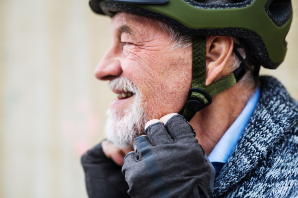 A close-up of active senior man with beard and mustache standing outdoors, putting on bicycle helmet.