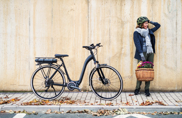 Active senior woman with electrobike and flowers in basket standing outdoors in town, leaning against a concrete wall. Copy space.