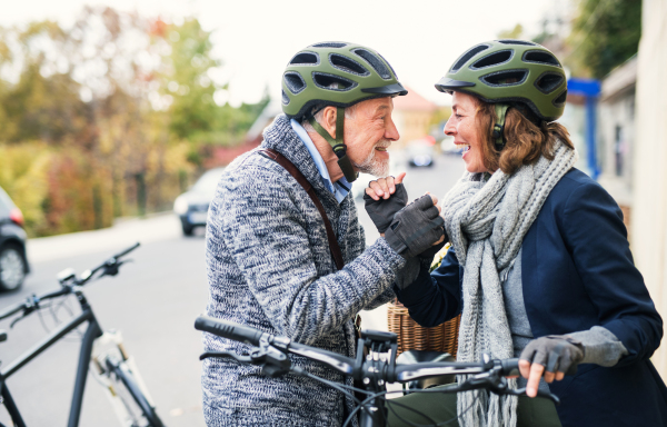 Active senior couple in love with helmets and electrobikes standing outdoors on a road in town, looking at each other.