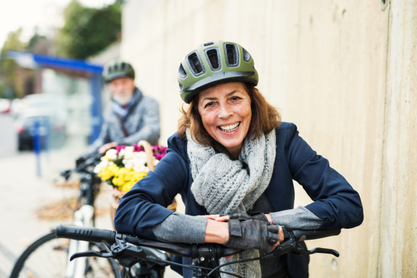 Active senior couple with helmets and electrobikes standing outdoors on a pathway in town.