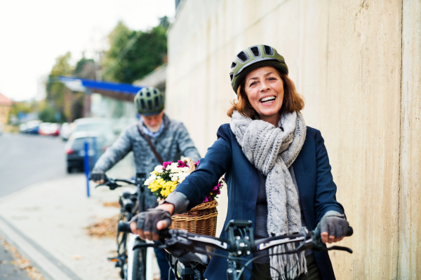 Active senior couple with helmets and electrobikes standing outdoors on a road in town.