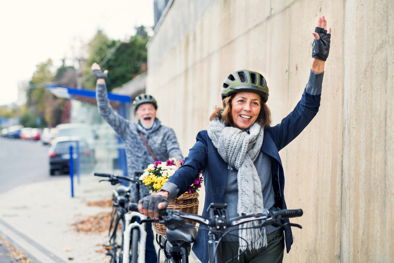 Active senior couple with helmets and electrobikes standing outdoors on a road in town, greeting somebody.