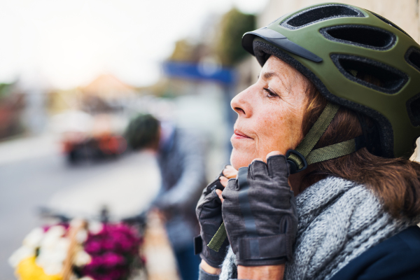 Active senior woman standing outdoors on a pathway in town, putting on a bike helmet. Copy space.