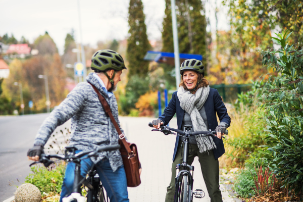 An active senior couple with helmets and electrobikes standing outdoors on a pathway in town, looking at each other.