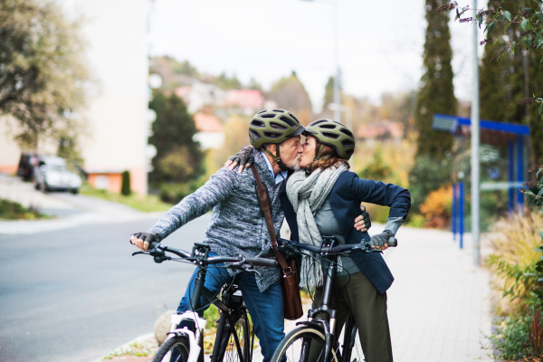 Active senior couple with helmets and electrobikes standing outdoors on a road in town, kissing.