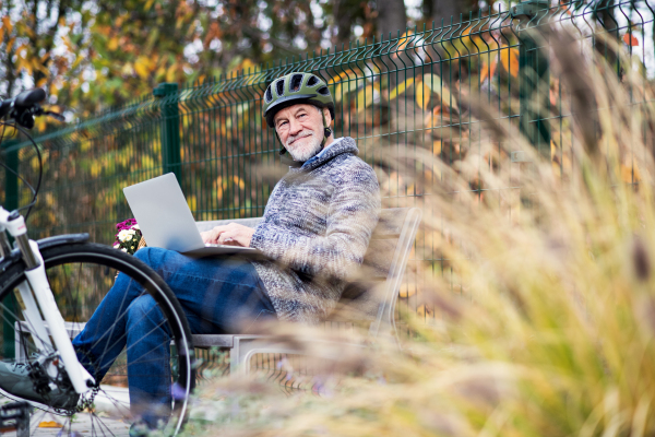 A senior man with electrobike sitting on a bench outdoors in town in autumn, using laptop. Copy space.