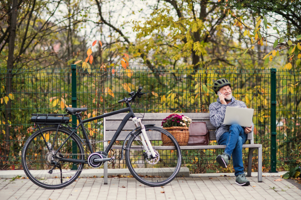 A senior man with electrobike, laptopand smartphone sitting on a bench outdoors in town in autumn, making a phone call. Copy space.