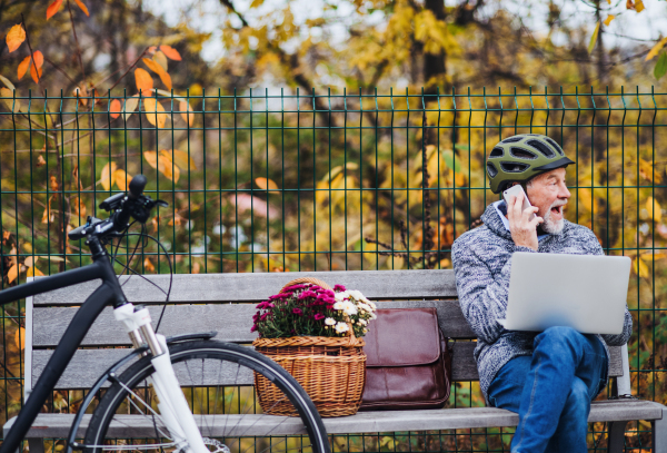 A senior man with electrobike, laptop and smartphone sitting on a bench outdoors in town in autum. Copy space.