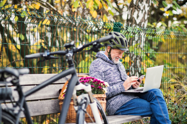 A senior man with electrobike, laptop and smartphone sitting on a bench outdoors in town in autum. Copy space.