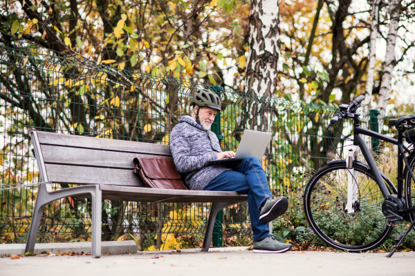 A senior man with electrobike sitting on a bench outdoors in town in autumn, using laptop. Copy space.