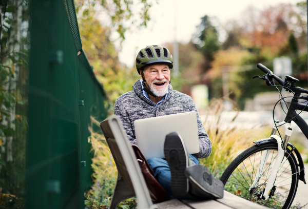 A senior man with electrobike sitting on a bench outdoors in town in autumn, using laptop. Copy space.