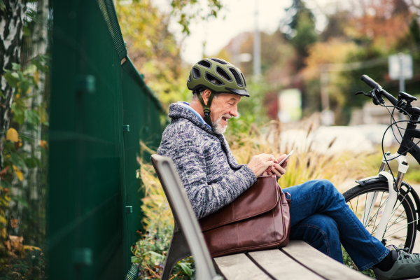 A senior man with electrobike sitting on a bench outdoors in town in autumn, using smartphone. Copy space.