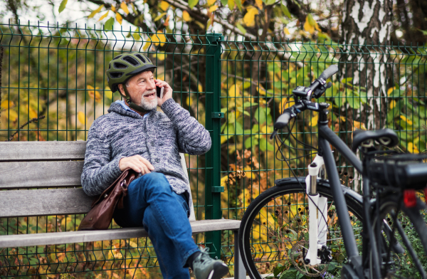 A senior man with electrobike and smartphone sitting on a bench outdoors in town in autumn, making a phone call. Copy space.