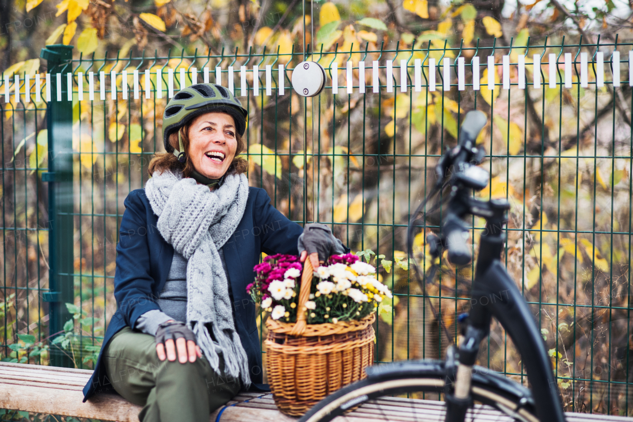 A senior woman with electrobike and flowers in a basket sitting on a bench outdoors in town in autumn. Copy space.