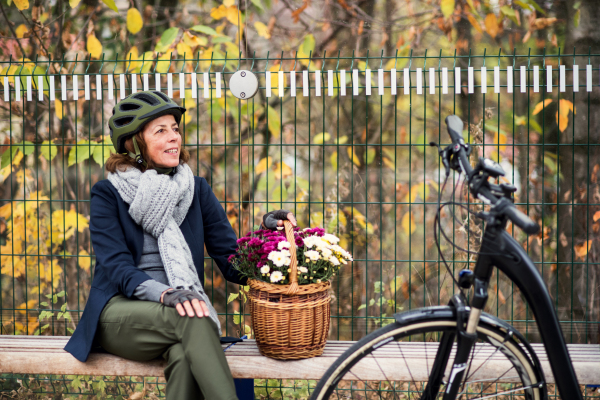 A senior woman with electrobike and flowers in a basket sitting on a bench outdoors in town in autumn. Copy space.