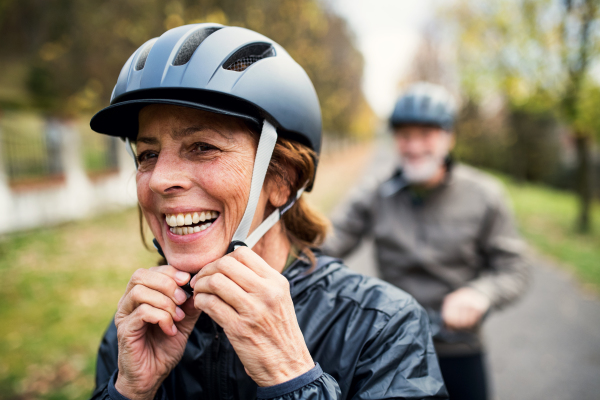 An active senior couple with helmets and electrobikes standing outdoors on a road in nature, putting on a helmet.