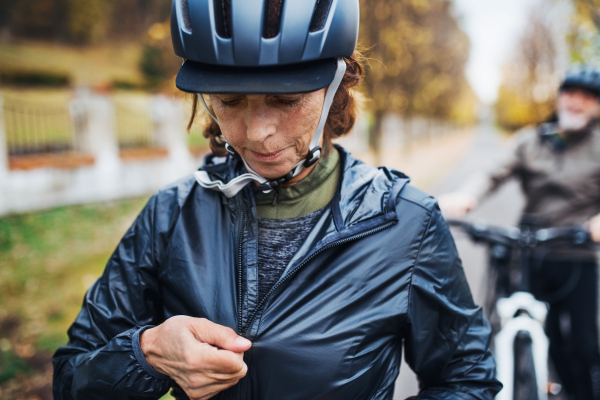 An active senior couple with helmets and electrobikes standing outdoors on a road in park.