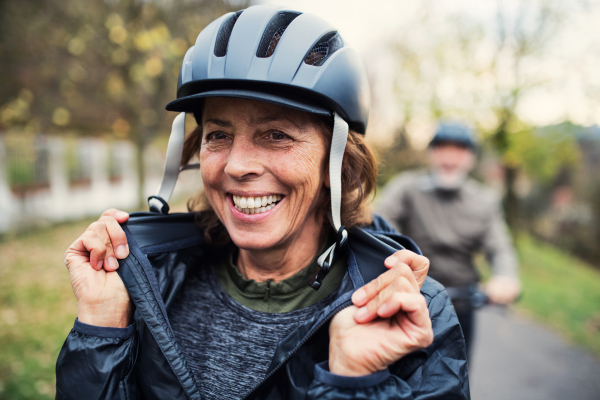 An active senior couple with helmets and electrobikes standing outdoors on a road in park.