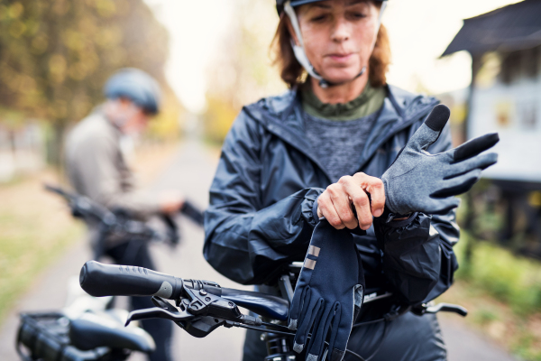 An active senior couple with helmets and electrobikes standing outdoors on a road in nature, putting on gloves.