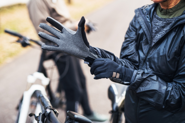 A midsection of active senior couple with electrobikes standing outdoors on a road, putting on gloves.