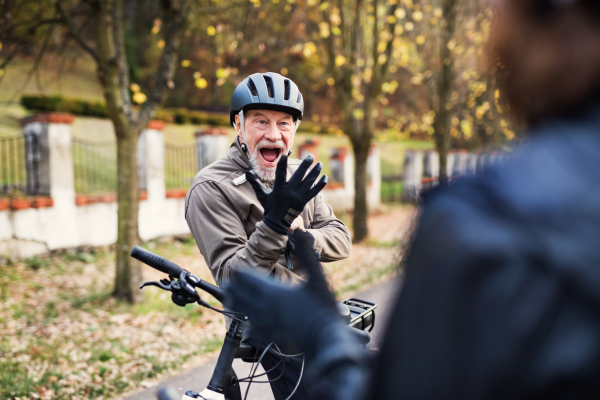 An active senior couple with helmets and electrobikes standing outdoors on a road in park, putting on gloves.