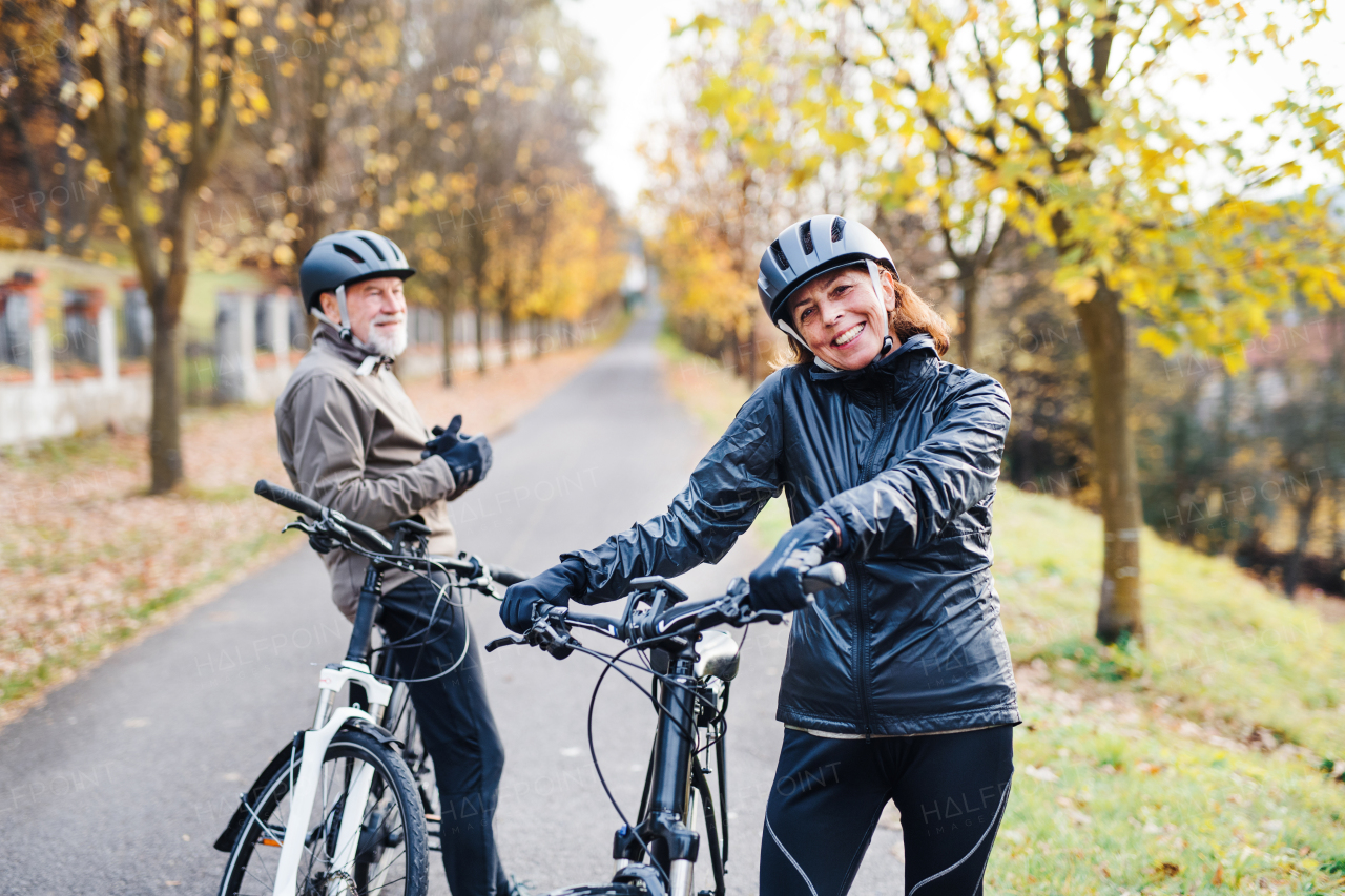 An active senior couple with helmets and electrobikes standing outdoors on a road in nature.