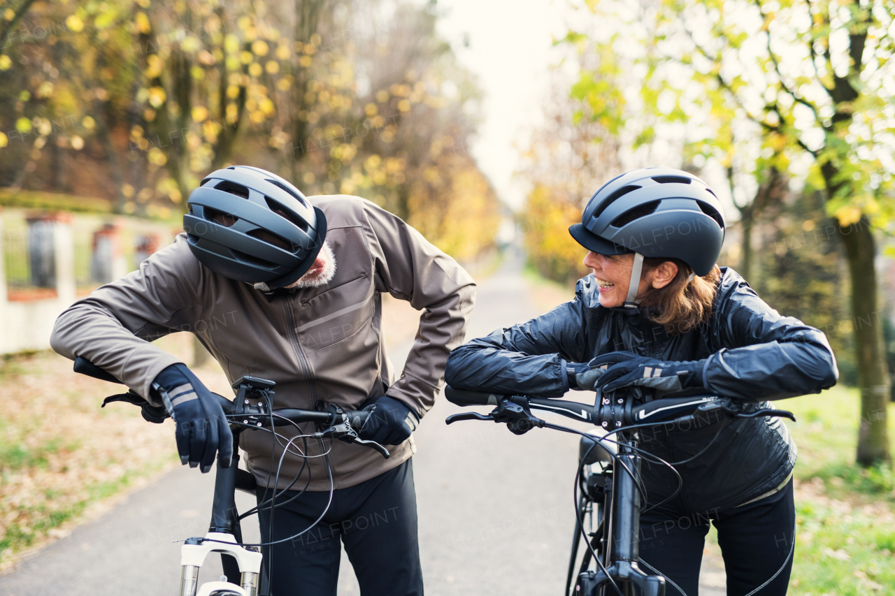 An active senior couple with helmets and electrobikes standing outdoors on a road in nature, looking at each other.