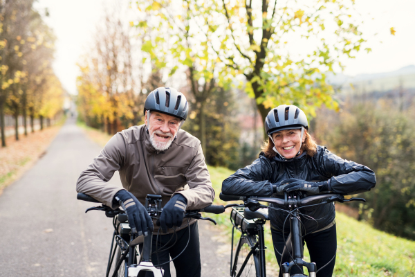 An active senior couple with helmets and electrobikes standing outdoors on a road in nature.