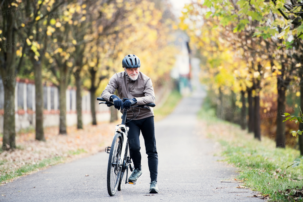 An active senior man with helmet and electrobike standing outdoors on a road in nature.