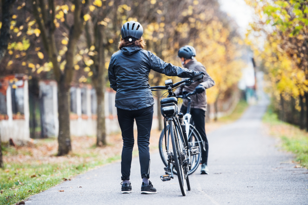 A rear view of active senior couple with helmets and electrobikes standing outdoors on a road in park.