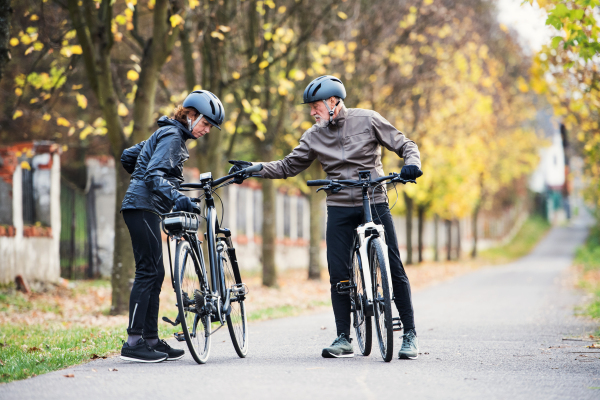 Active senior couple with helmets and electrobikes standing outdoors on a road in nature, talking.