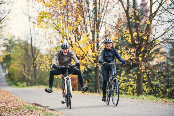 An active senior couple with helmets and electrobikes cycling outdoors on a road in nature, having fun.