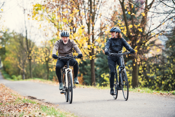 An active senior couple with electrobikes cycling outdoors on a road in park in autumn.