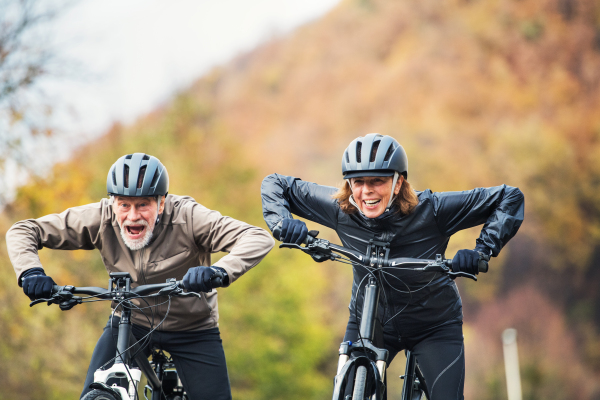 An active senior couple with helmets and electrobikes cycling outdoors on a road in nature.