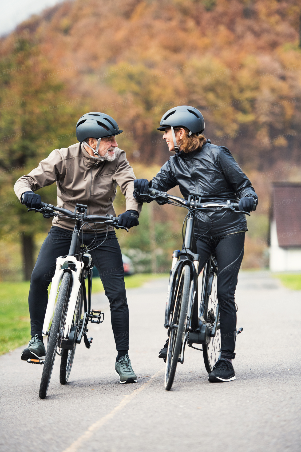 An active senior couple with helmets and electrobikes standing outdoors on a road in nature.