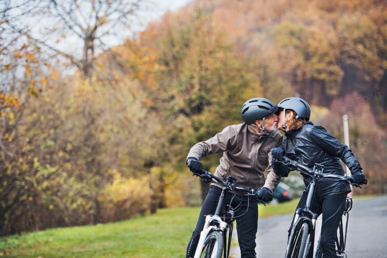 An active senior couple in love with helmets and electrobikes standing outdoors on a road in nature, kissing. Copy space.