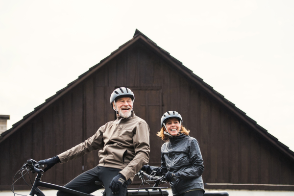 An active senior couple with helmets and electrobikes standing outdoors in front of a house.