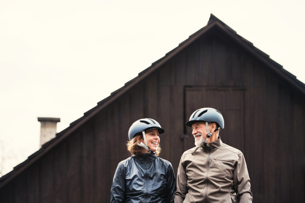 Happy active senior couple with bike helmets standing outdoors in front of a house.