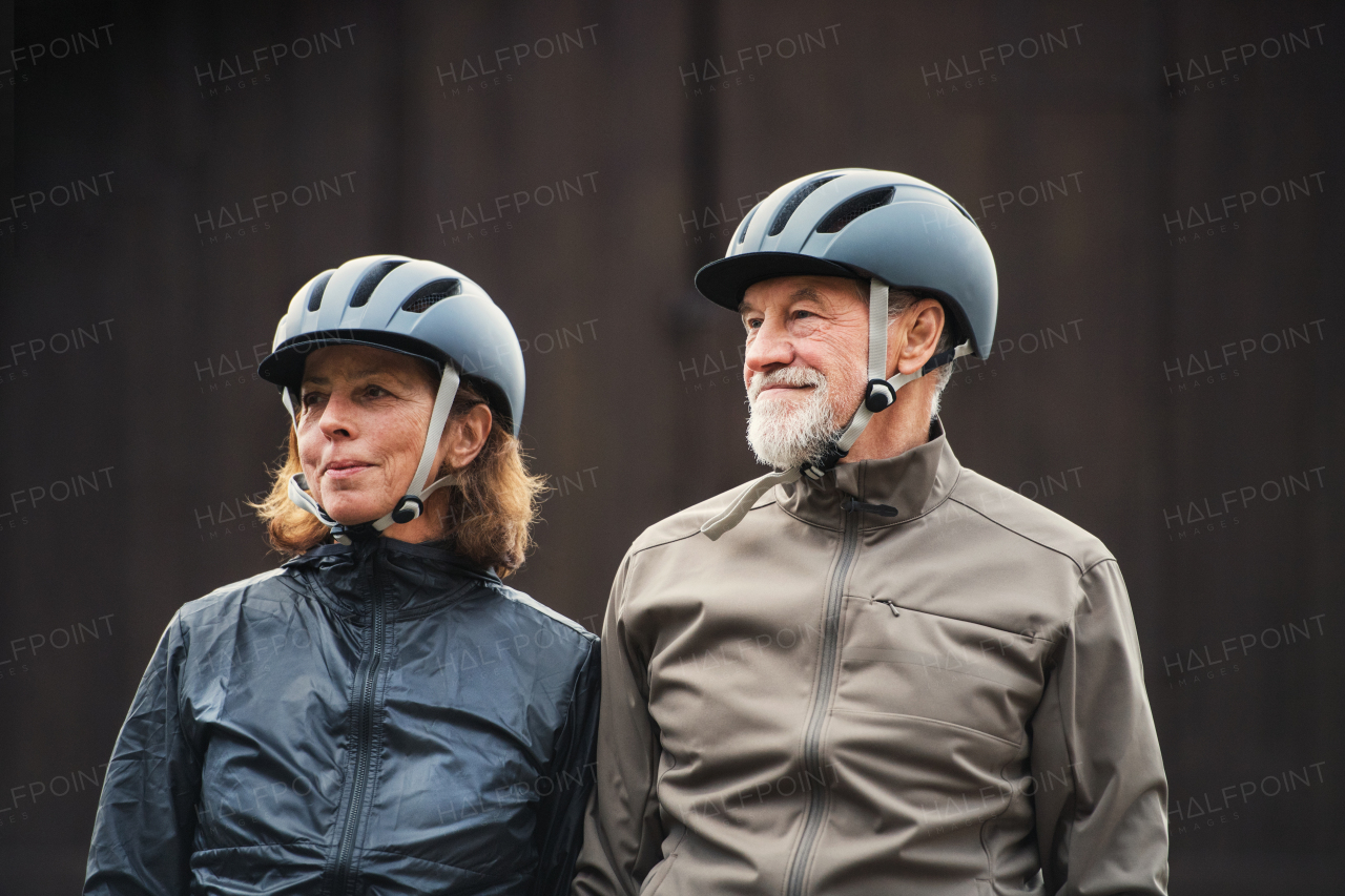 Happy active senior couple with bike helmets standing outdoors against dark background.