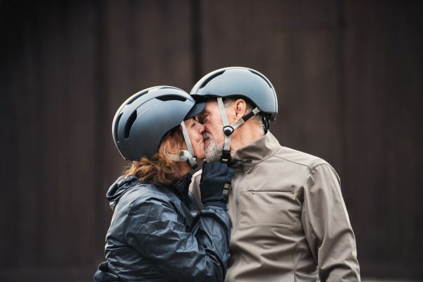 Happy active senior couple with bike helmets standing outdoors against dark background, kissing.