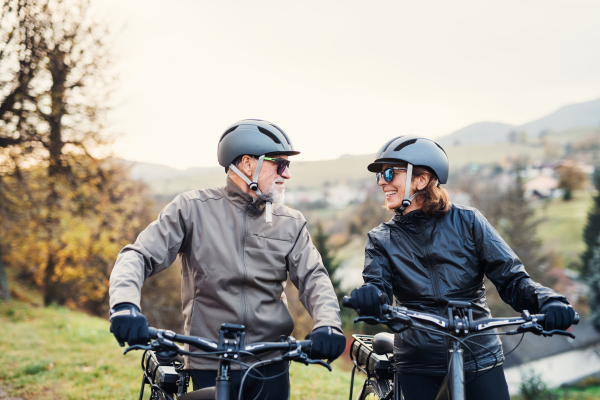 An active senior couple with helmets and electrobikes standing outdoors on a road in nature.