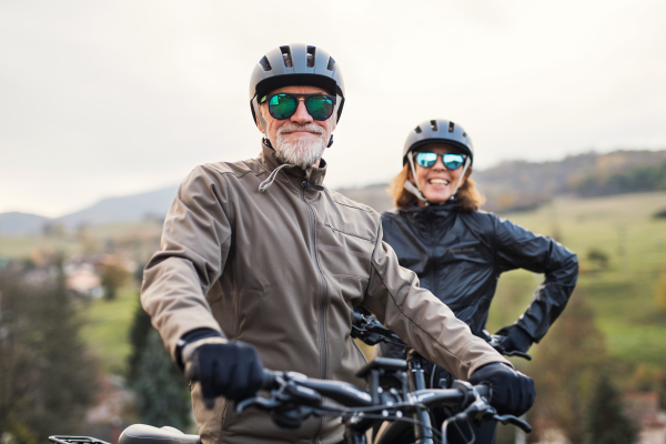 An active senior couple with helmets and electrobikes standing outdoors on a road in nature.