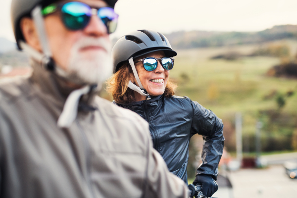 An active senior couple with helmets and electrobikes standing outdoors on a road in nature.