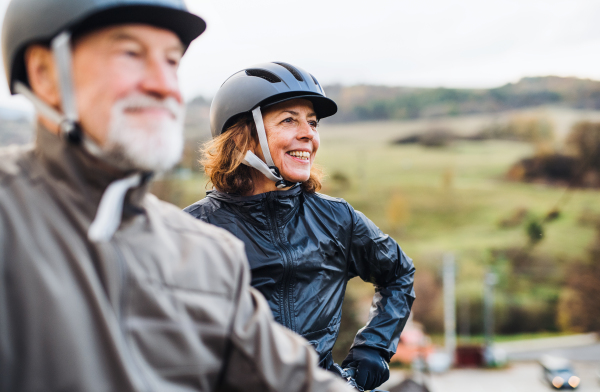 An active senior couple with helmets and electrobikes standing outdoors on a road in nature. Copy space.