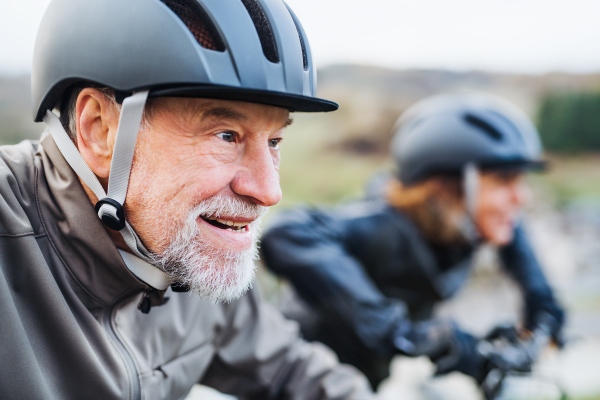 An active senior couple with helmets and electrobikes cycling outdoors on a road in nature.