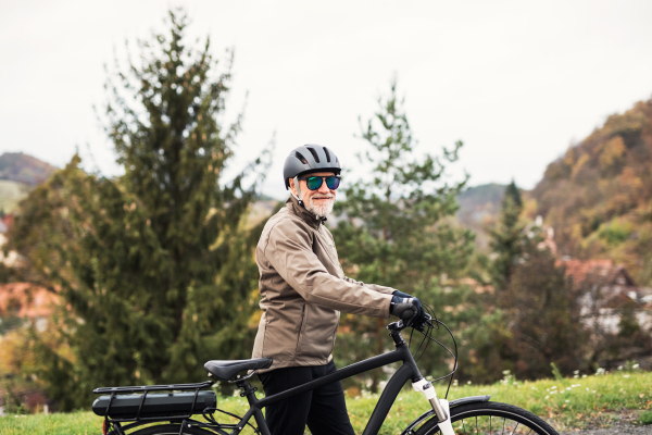 An active senior man with helmet, sunglasses and electrobike standing outdoors on a road in nature.