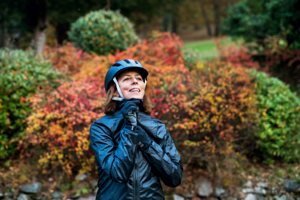 Active senior woman with sports jacket and gloves standing outdoors on a road in nature, putting on bicycle helmet. Copy space.