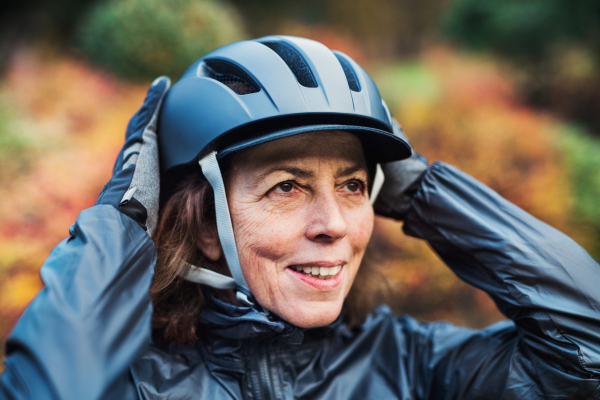 A close-up of active senior woman standing outdoors in nature, putting on bicycle helmet.