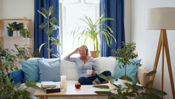 Happy attractive senior woman with headphones sitting indoors on sofa, relaxing.