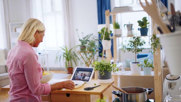 Attractive senior woman preparing food in kitchen indoors, following food vlogger on tablet.